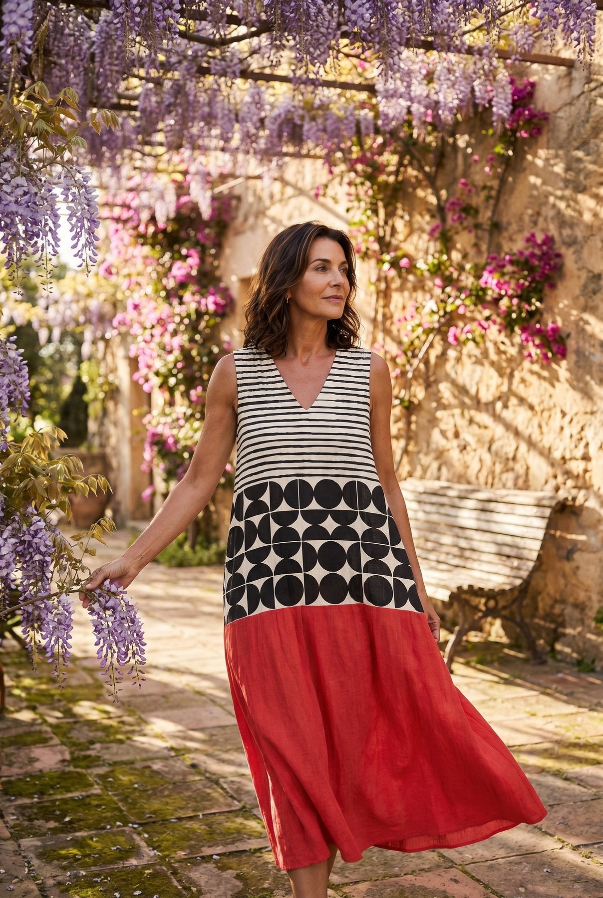 The Mary Boat Dress in stripes-and-dots, editorial full-body in a wisteria pergola courtyard with cascading purple blooms