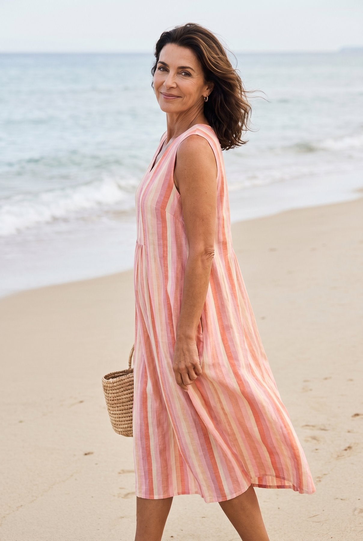The Nancy Print Dress in pink and coral vertical stripes, sleeveless V-neck midi, full-body editorial on a Mediterranean beach with woven basket